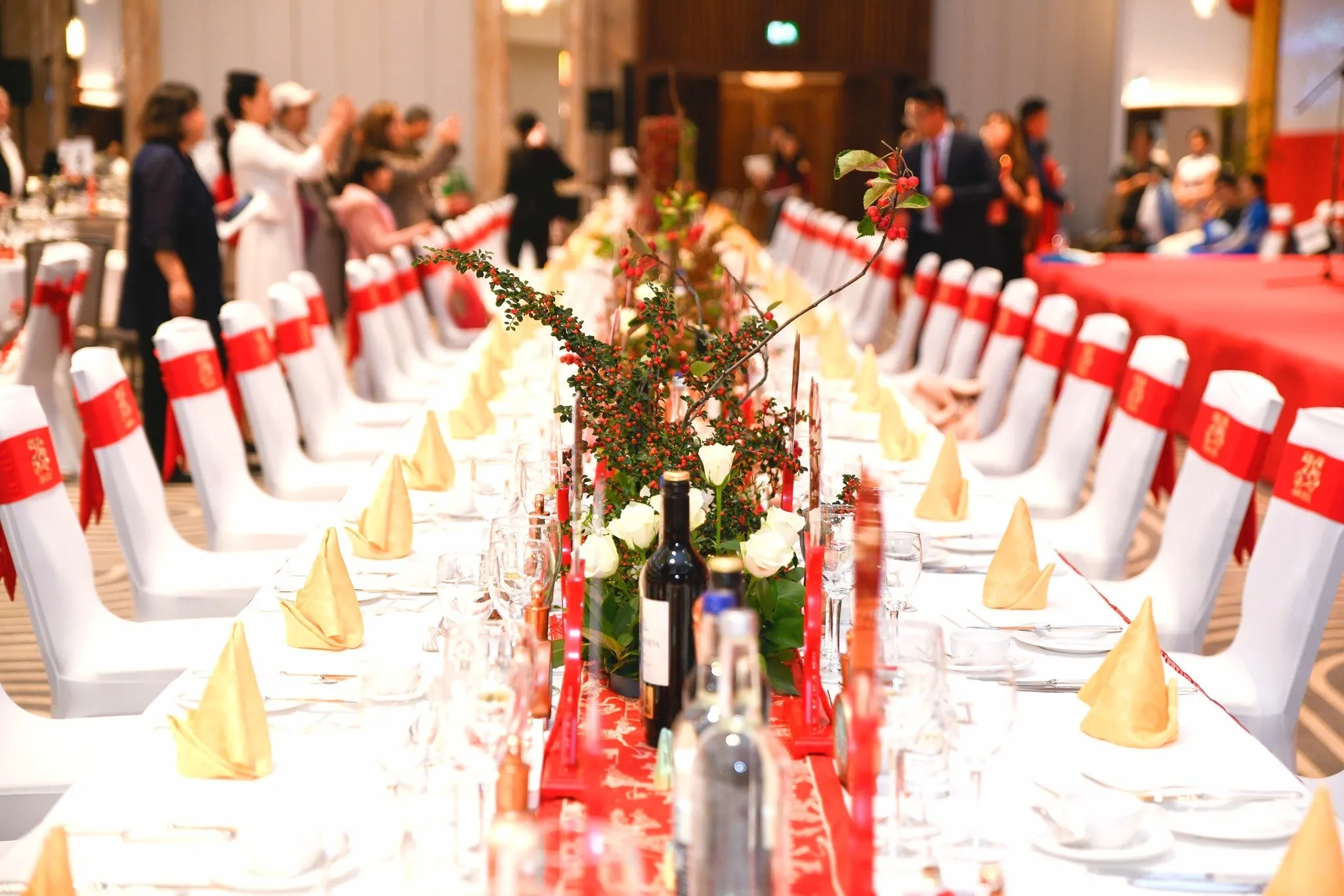A long banquet table with white and red chair covers, folded gold napkins and a berry centrepiece, guests gathering beyond